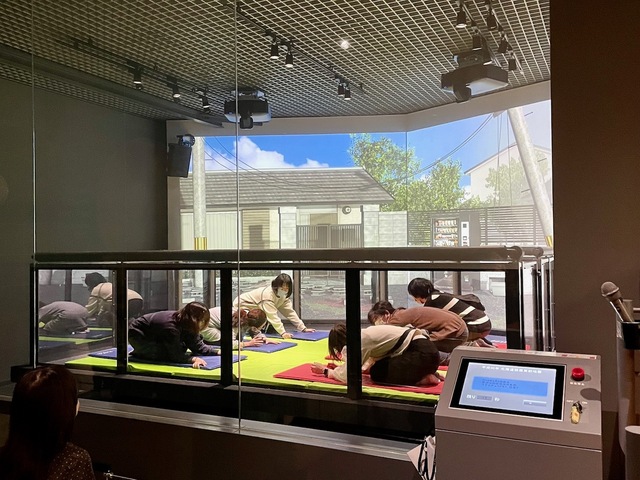 【本所防災館】地震体験をする小学生。スクリーンには地震で倒れる電柱などが映し出されます