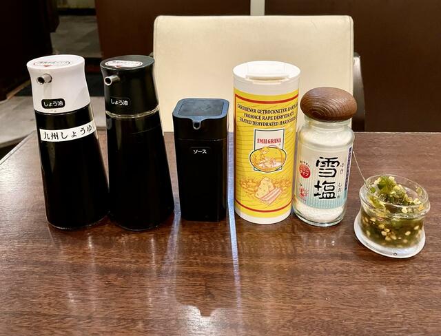 【トーキョーアジフライ】各テーブルやカウンターに用意された調味料。黄色い筒が粉チーズ。右が今月の薬味、青唐辛子の醤油漬け