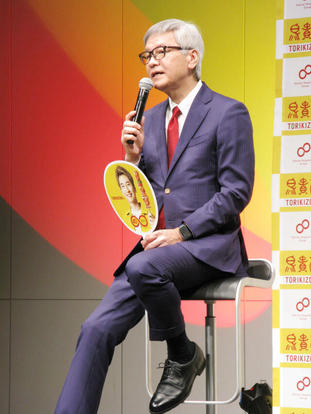 息子の笑顔がプリントされたうちわを手に語る大倉社長。