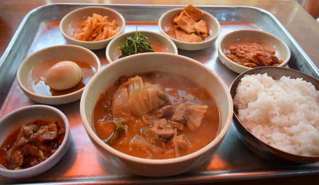 『スリョンチプ』のキムチチゲ定食4000ウォン。手前中央のキムチチゲから時計回りに、貝の塩辛、茹で卵、切り干しダイコンのナムル、青菜のナムル、オデン（魚のすり身）、ハクサイキムチ、大盛りごはん。この内容で4000ウォンを維持しているとはまったく頭が下がる