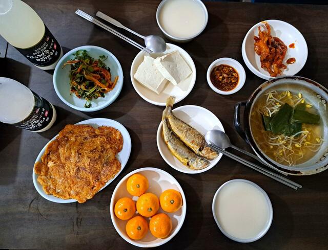 【美食と美酒の都、全州特集 vol.3】西部市場近くの酒場のおまかせつまみ。上段左から全州生マッコリ、切り干し大根とパクチーのナムル、豆腐、鶏の足の辛煮。下段左からキムチのジョン、イシモチの揚げ焼き、豆モヤシのスープ、みかん