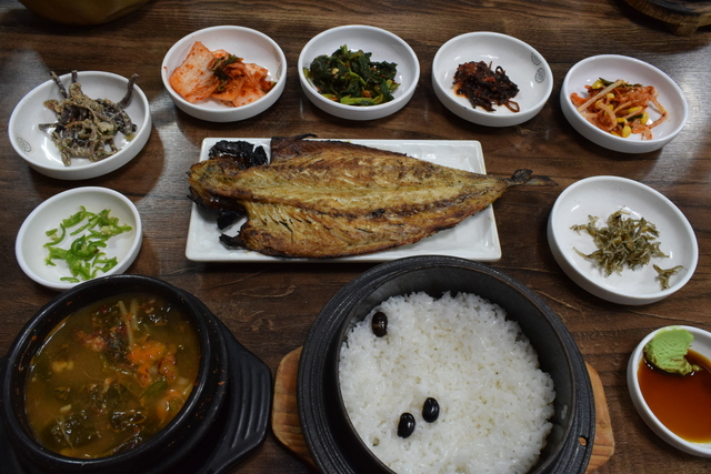 『全州食堂』のコドゥンオ（サバ）定食9000ウォン。手前のごはん（黒豆入り）から時計回りに、チゲ、青唐辛子（お好みでチゲに入れる）、ワラビのナムル、ハクサイキチム、ホウレンソウのナムル、葉トウガラシのナムル、豆モヤシのナムル、ジャコ炒め、ワサビ醤油（焼サバをつけて食べる）。中央が焼サバ
