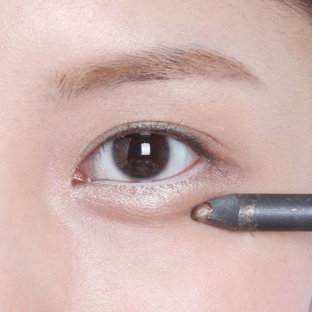 5. Dで涙袋の下にラインを描く／下まぶたに沿って涙袋を強調する。成功の秘密はふたつ。1. 微笑んで涙袋が目立つようにする! ２. 写真のようにペンシル本体が顔につくように横にして描く! 