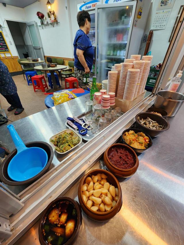 以前のように店の外にカウンターはない。入口が二重になっていて、入ってすぐのところにあるカウンターに食べ放題のつまみが並んでいる。右から煮干し、大根のキムチ、味噌ダレ、ニンニク、醤油ダレ。栓抜きの隣はチヂミ。つまみの種類は半減したが、物価高、とくに野菜の高騰などを考えるとやむをえないことだろう